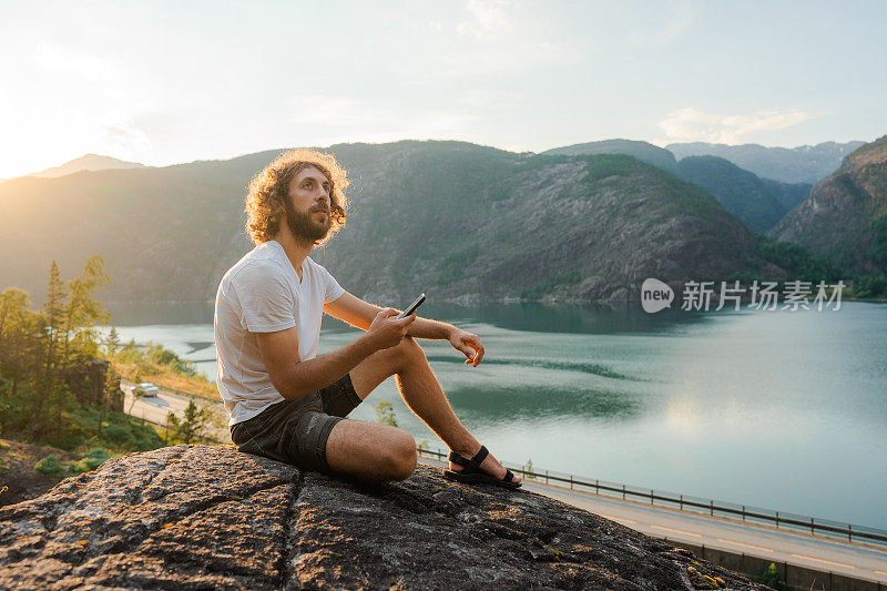 一名男子一边坐着一边玩智能手机，一边欣赏挪威峡湾的景色