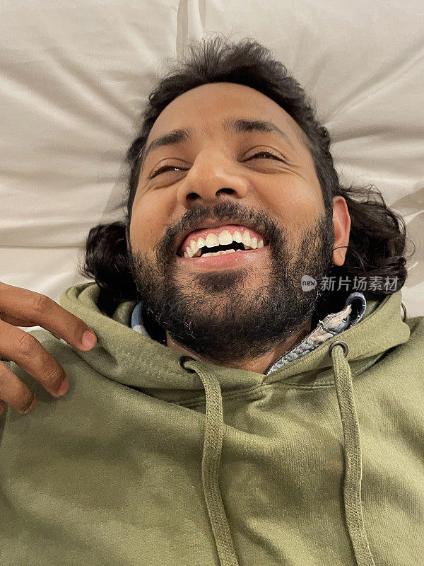 全画幅照片，印度男子仰面倒在酒店房间的双人床羽绒被上，仰面躺着笑，看着相机，高架视图