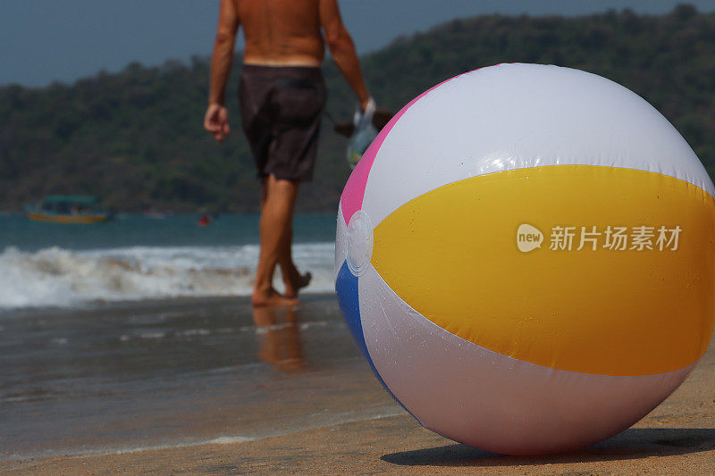 水边沙滩上的沙滩球的特写图像，塑料，色彩鲜艳的儿童海滩玩具，退潮时无法辨认的游客走过破碎的海浪，林地背景，重点是前景