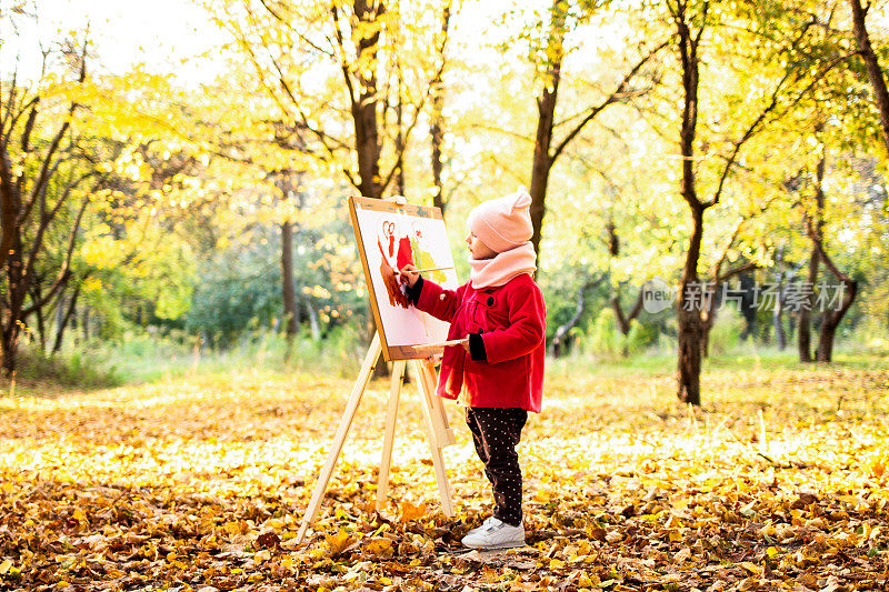 一个穿着红色外套的小女孩在公园的画架上画着一幅水彩画，背景是秋天的风景