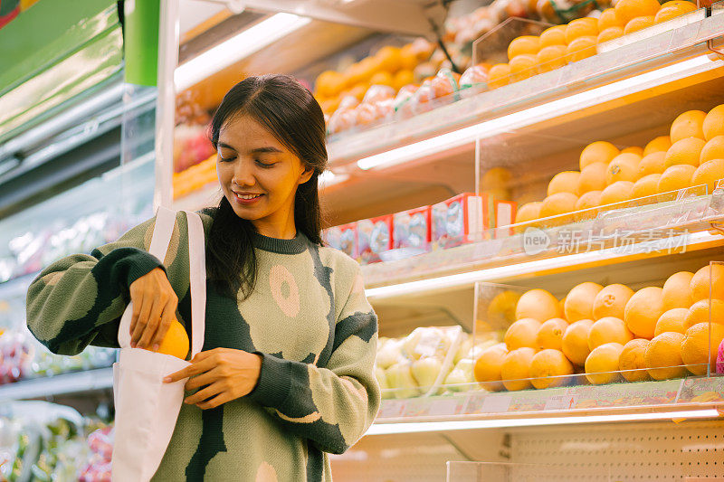 一名亚洲妇女用可重复使用的袋子在杂货店购物时拿着橙子。