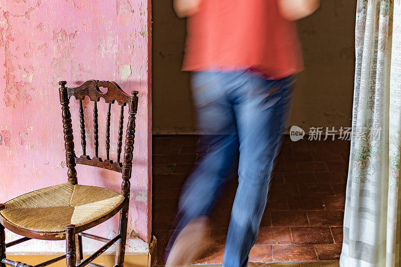 一个男人转身走进一间饱经风霜的老房子。他的身影因运动而模糊不清。一张古色古香的椅子在前景中占据突出位置。