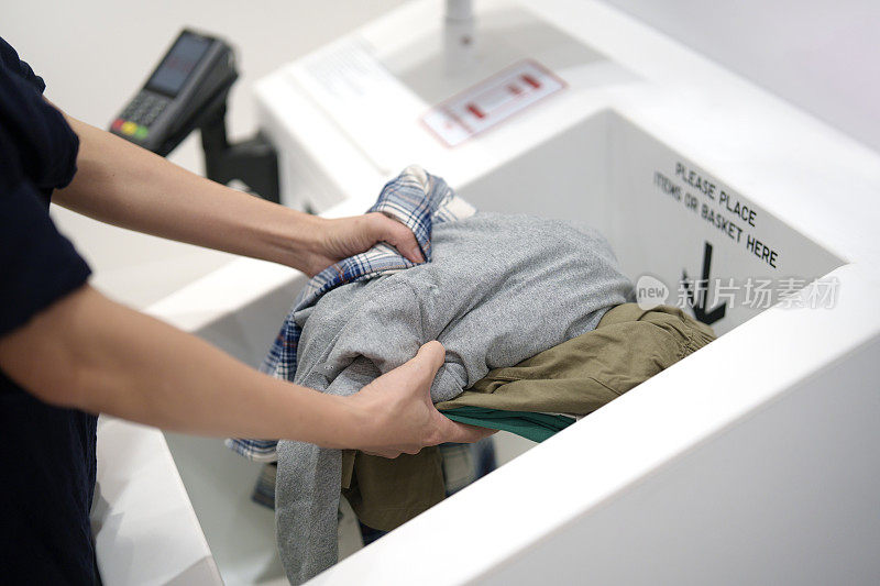 一名亚洲妇女在使用自助结账柜台和非接触式支付系统前，将购物衣物放入RFID自助结账系统