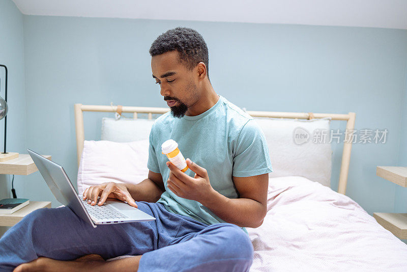 年轻的非裔美国人在卧室里用笔记本电脑，拿着药瓶