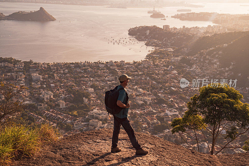 在黄金时段，一个孤独的徒步旅行者站在山顶上，凝视着城市令人惊叹的风景。太阳在远处落下，温暖的光芒笼罩着这座城市及其标志性的多山海岸线。