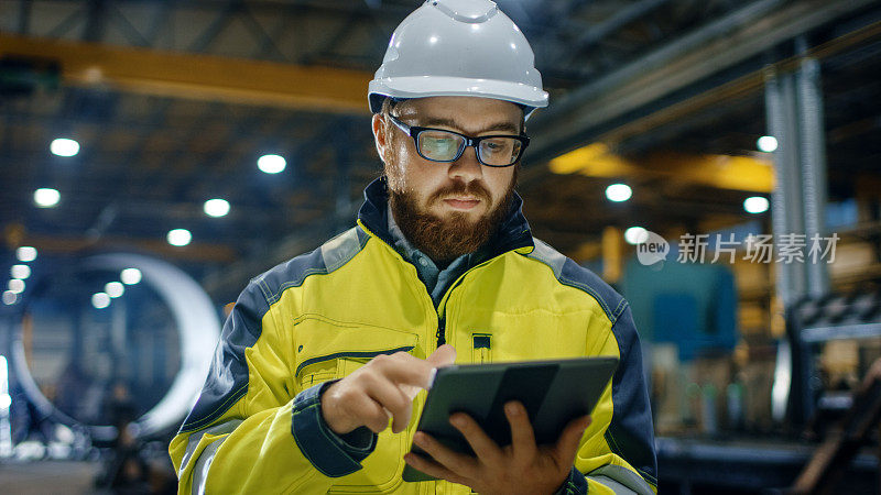 工业工程师戴安全帽穿安全服使用触屏平板电脑。他在重工业制造厂工作。