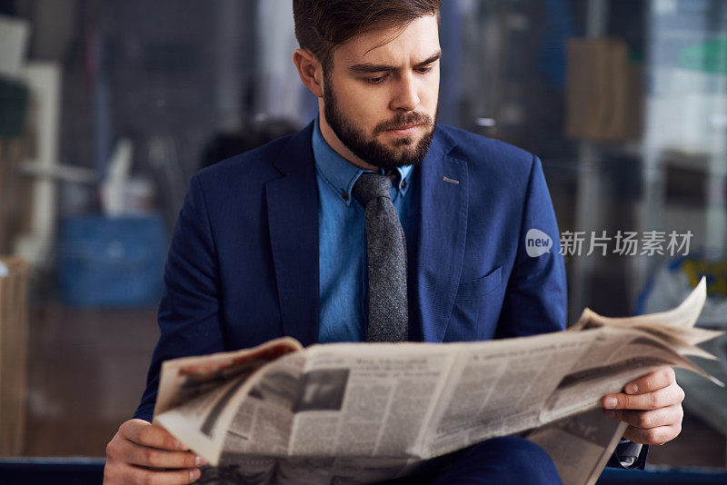 穿着蓝色西装、蓄着胡须、神情凝重的英俊年轻男子坐在机场里读着最新的新闻来浪费时间