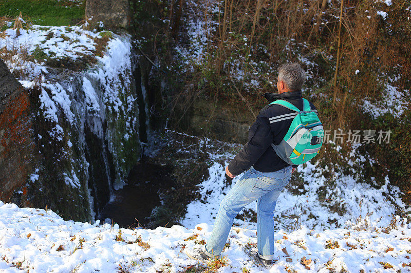 成年男子徒步旅行在大雪纷飞的冬季大自然旁边结冰的小瀑布