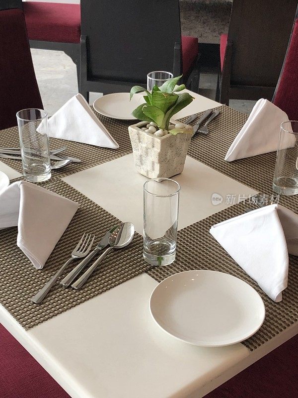 白色方形餐厅餐桌的形象，为早餐，午餐或晚餐照片与四个棕色的餐垫，银餐具刀，叉和勺子，盘子，水杯，米色和白色主题折叠布餐巾，红色椅子
