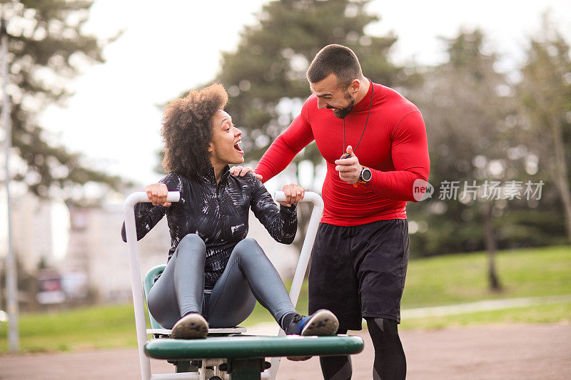 肌肉发达的私人教练鼓励一名女运动员在城市公园的划船机上锻炼，并在秒表上显示她的时间