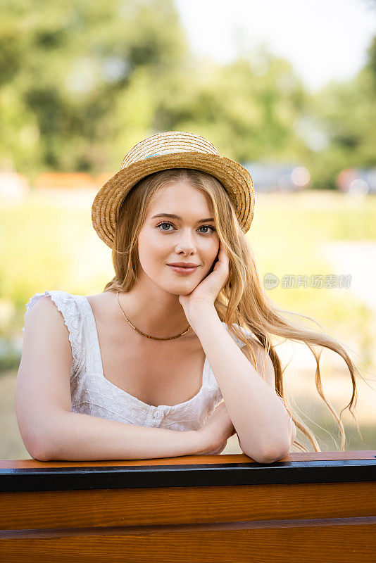 漂亮的女孩戴着草帽，坐在长椅上看着镜头
