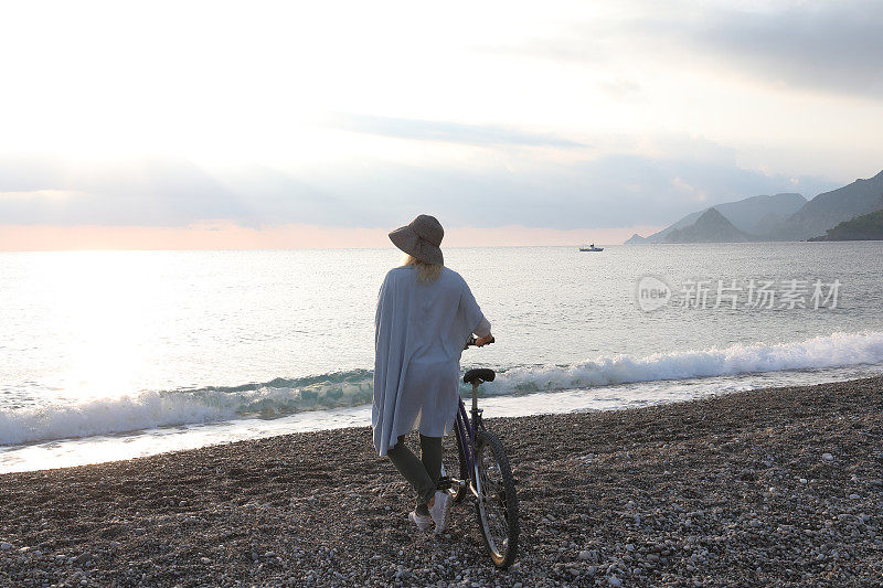 成熟的女人骑着自行车在日出的海滩上放松