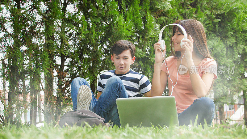 人们拥有现代科技。一对大学朋友坐在公园里。亚洲女孩戴着耳机在网上上课