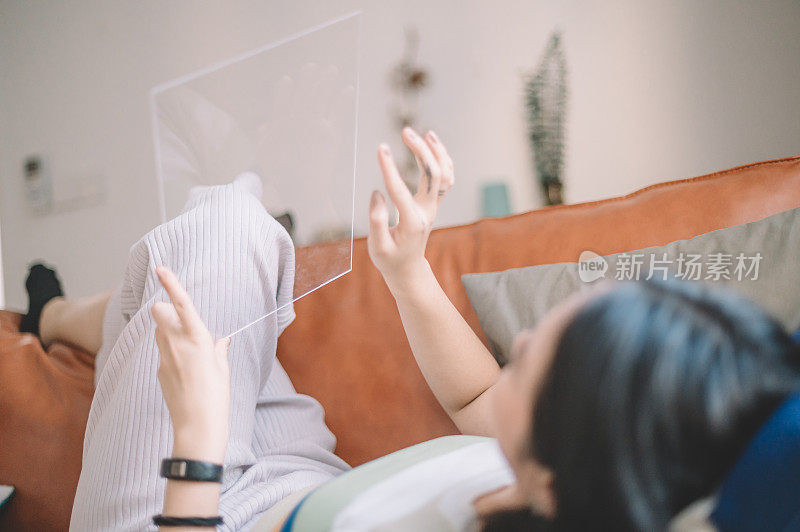 一名亚裔华裔女大学生躺在沙发上用自己的透明数字平板电脑与朋友聊天