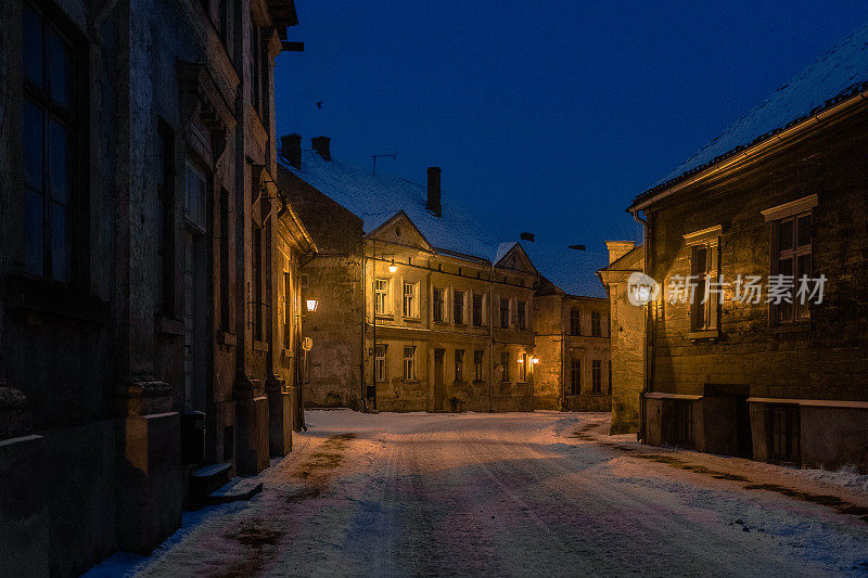 雪覆盖的街道Kuldiga老城在蓝色的时间照明路灯。
