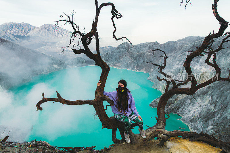 印度尼西亚东爪哇岛，一名女子坐在一棵枯树上，观看伊真火山火山口和山脉中美丽的绿松石湖