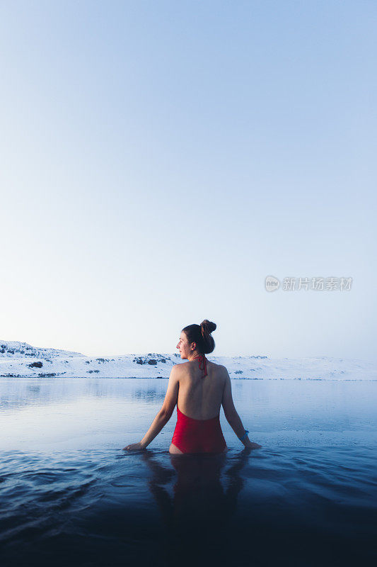 一名女子正在温泉池中沐浴，可以看到冰岛的雪山和冰湖