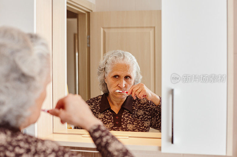 越过肩膀的观点，一个美丽的老妇人与灰色的头发刷牙，看着自己在浴室的镜子和水槽
