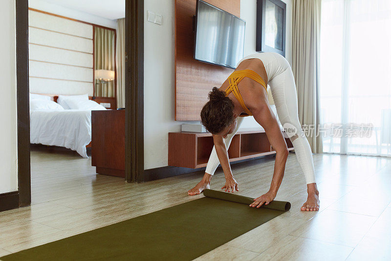 培训在家里。女人在客厅练习瑜伽后，滚动瑜伽垫。女性在完成日常运动后，在运动服装折叠健身地毯。在社会隔离期间保持活跃和健康。
