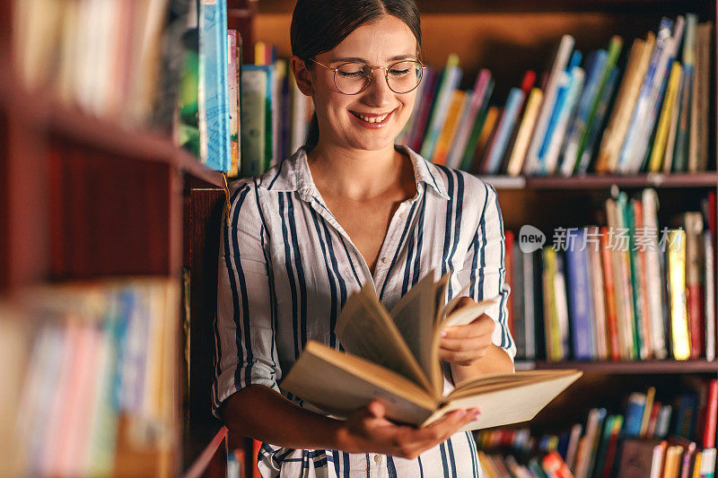 年轻迷人的女大学生倚在图书馆的书架上看书。