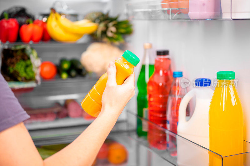一个女孩拿着一瓶橙色的蔬菜或果汁，背景是一个打开的冰箱和食物。饮食运动健康饮品概念，近景，浅景深。