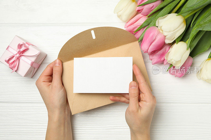 女性手拿信封与空白卡片白色木质背景与郁金香和礼盒