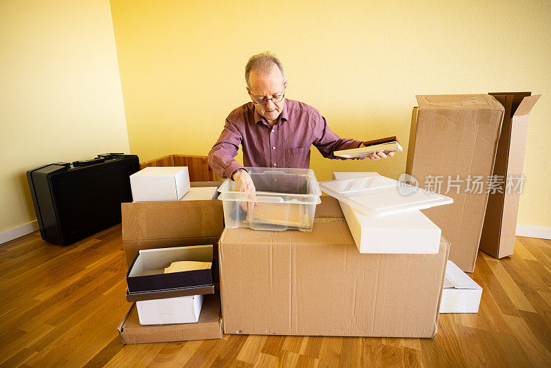 一个年长的白人男子在空荡荡的公寓房间里查看箱子里的东西，他正在从纸箱里取出要搬家的东西