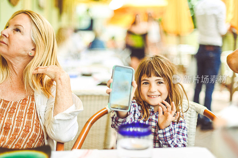 孩子在户外餐厅和她的祖母和家人一起使用手机，坦诚的家庭团聚