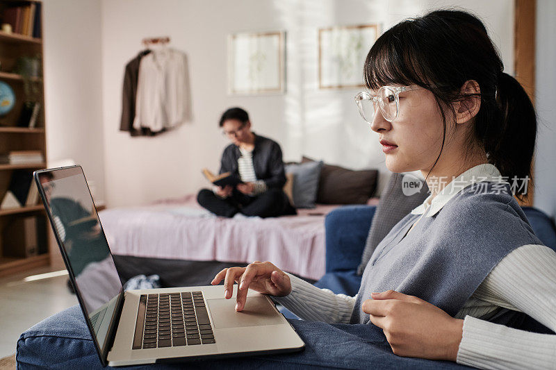 全神贯注的亚洲学生女孩戴着眼镜在笔记本电脑上搜索信息，而她的室友看书