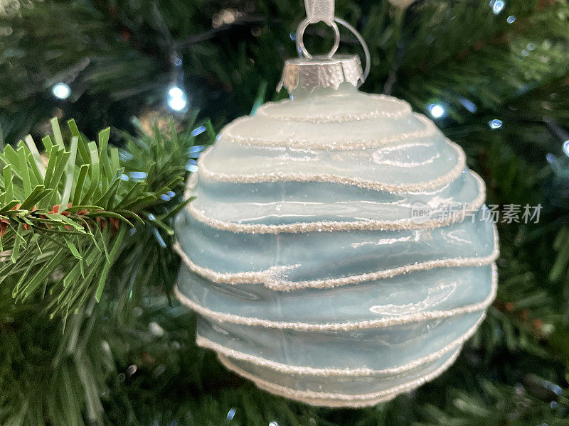 个人的特写图像，蓝色的肋小装饰物挂在人造圣诞树的树枝上，云杉针，模糊的白色仙女光的背景