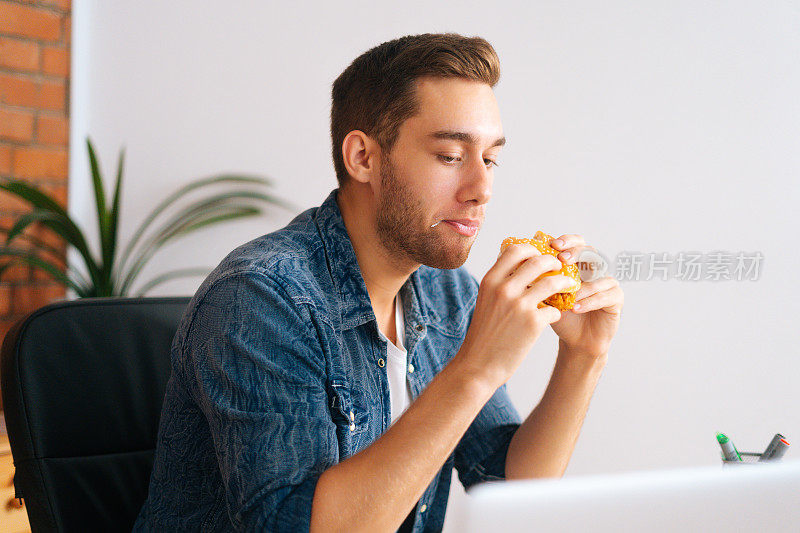 侧视图英俊的年轻人吃汉堡看在线电影笔记本电脑坐在办公桌在家庭办公室。