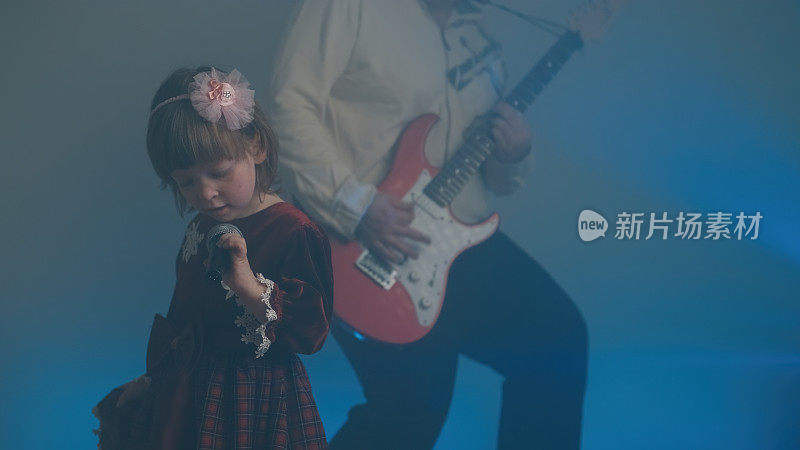 穿着复古礼服的小女孩在舞台上唱歌，她的父亲在弹奏电吉他