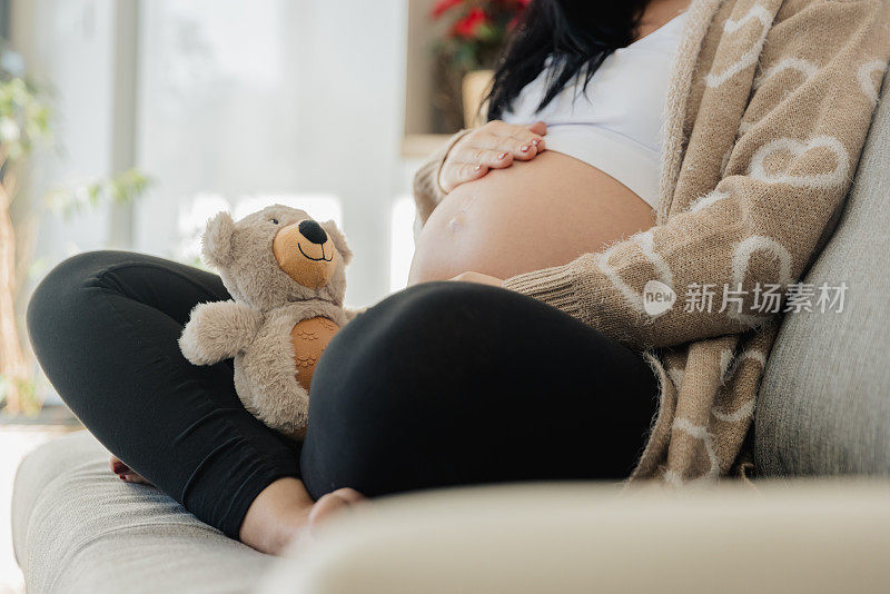 抱着泰迪熊的孕妇坐在沙发上摸肚子