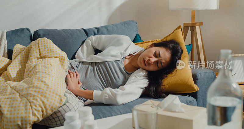 生病的年轻亚洲妇女遭受强烈的腹部疼痛躺在毯子下的沙发在家里的客厅。