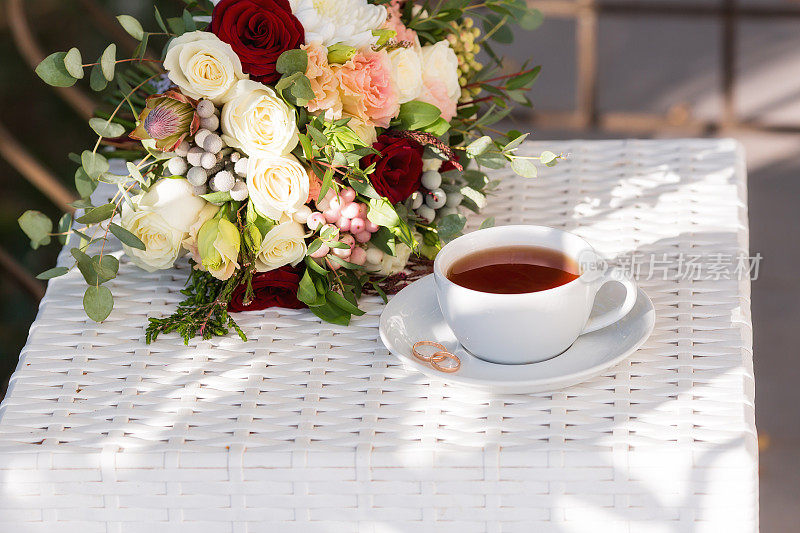 一杯茶，玫瑰花瓣，金婚戒指和一束美丽的花束放在白色的桌子上。婚礼庆典的概念。新娘的早安。