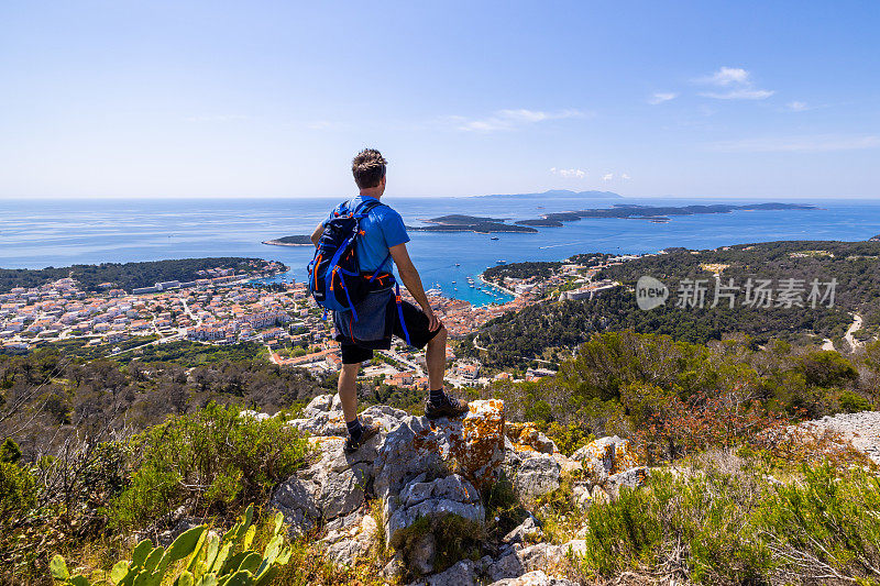 背着双肩包的男子站在岩石上，从山顶欣赏小镇的景色