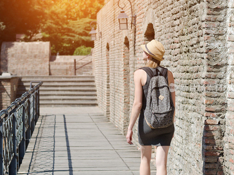 戴着帽子背着背包的女孩沿着城墙走着。阳光灿烂的日子