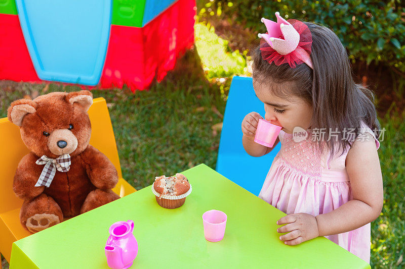 蹒跚学步的小女孩在户外茶会上玩，喝着杯子里的东西，最好的朋友泰迪熊坐在桌边。