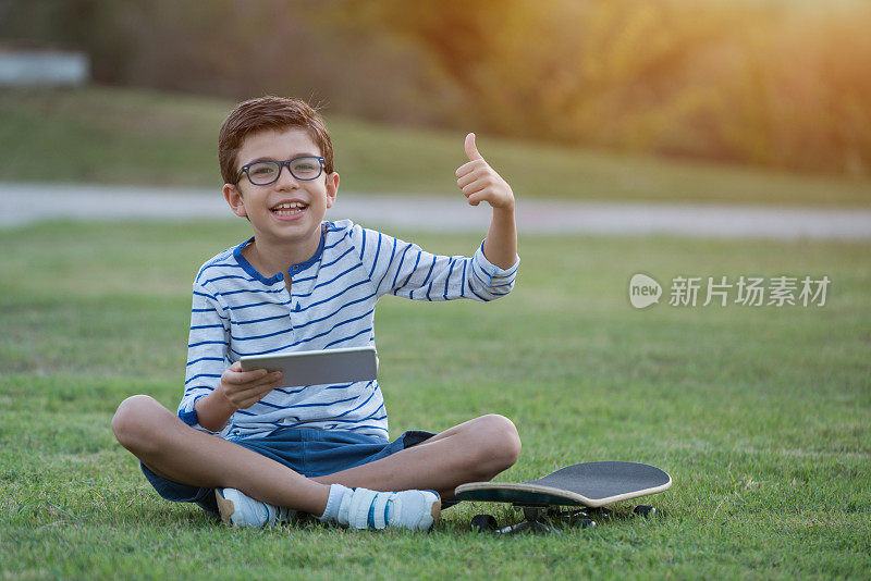 拿着平板电脑和滑板的男孩坐在草地上