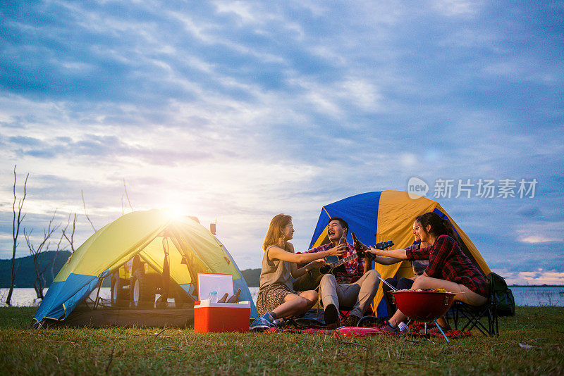 一群男人和女人喜欢在湖边露营野餐和烧烤，背景是帐篷。年轻的混血亚裔男女。年轻人的手为啤酒瓶干杯欢呼。