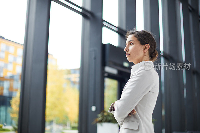 认真自信，有吸引力的年轻混血商务女士站在现代的大厅大公司，保持双臂交叉，而环顾四周