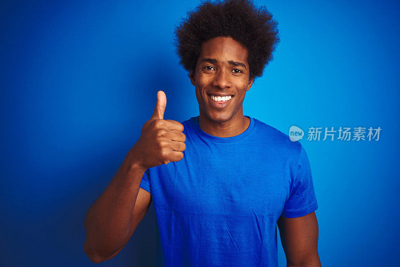 非洲裔美国人与非洲发式穿t恤站在孤立的蓝色背景做快乐的拇指手势与手。赞许的表情看着镜头，表示成功。