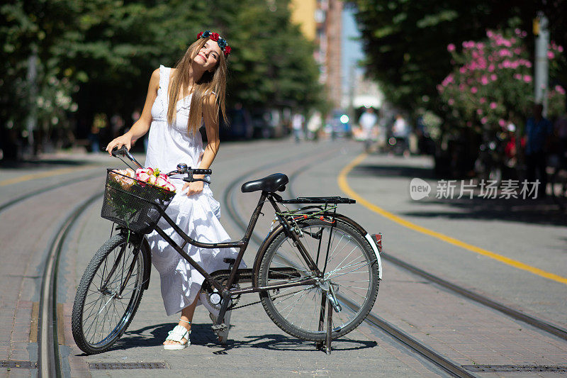 美丽的女自行车手在白色连衣裙停了一会儿，看着远处，而骑着她的蓝色自行车在城市中心与绿色的树的背景。美女集中放松夏天