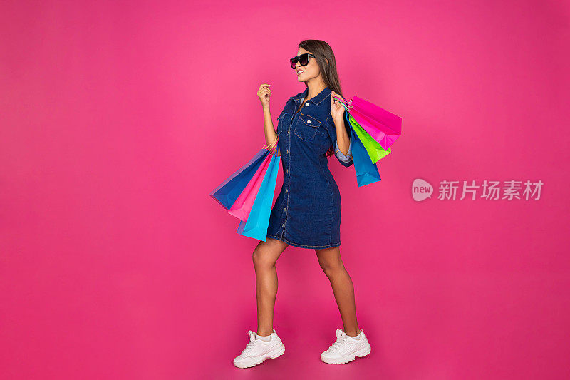 年轻美丽的女孩在牛仔裙携带购物袋在粉红色的背景。购物概念，夏季大甩卖。