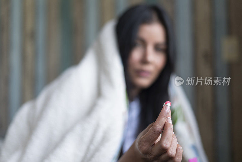 由于在床上隔离期间发高烧，年轻的拉丁裔女孩裹着毯子，她看着照片中的相机，手里拿着药片。