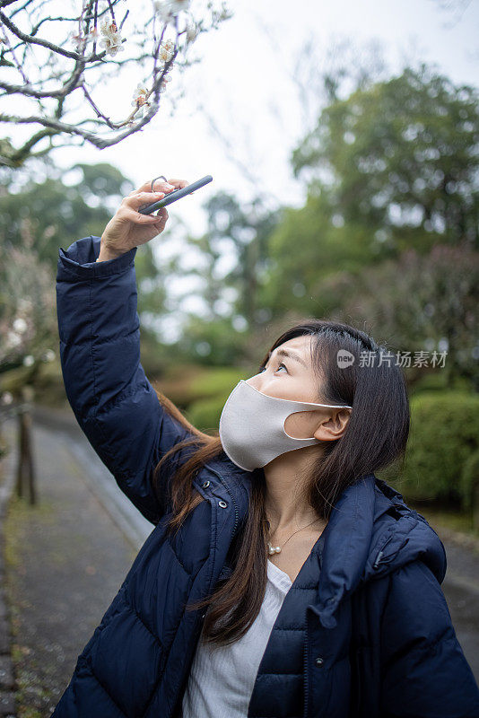 年轻女子戴着防护面罩在公园里拍摄梅花的照片