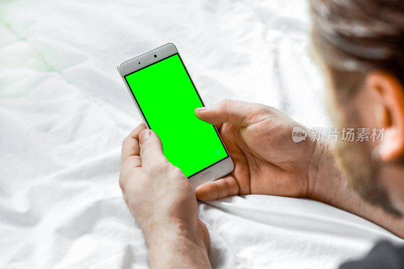 年轻的大胡子男子使用绿色色度键屏幕手机，放松在床上。有人使用绿色模拟屏幕智能手机。后面的观点。男子躺在白色毯子上，在ChromaKey显示器上观看视频