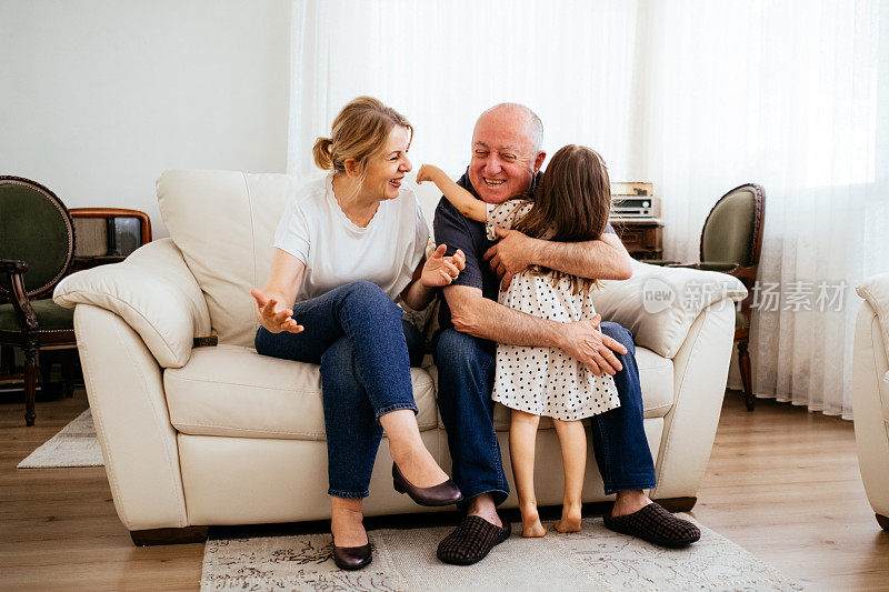 一家人在客厅里，爷爷奶奶和小女孩在玩，对着镜头微笑。幸福享受居家生活。奶奶坐在沙发上拥抱和亲吻可爱可爱的孩子。