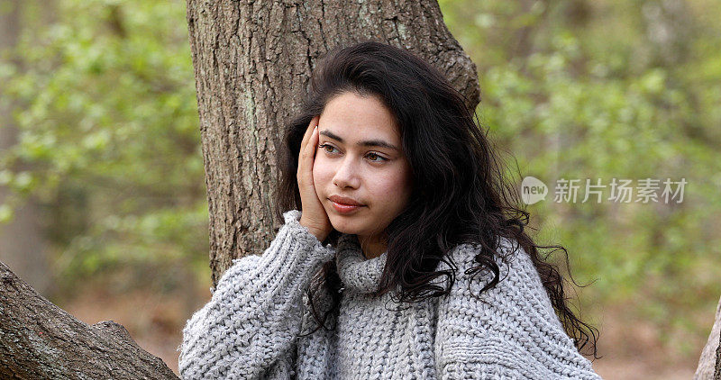 一个留着长发的年轻女子站在一棵树旁，向一边看。她穿着一件灰色的针织毛衣。