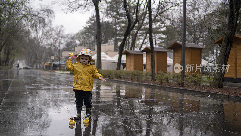 可爱调皮的小男孩，穿着亮黄色雨衣和胶靴，在春雨天潮湿的街道上和小水坑里的橡皮鸭玩耍。季节性天气步行概念